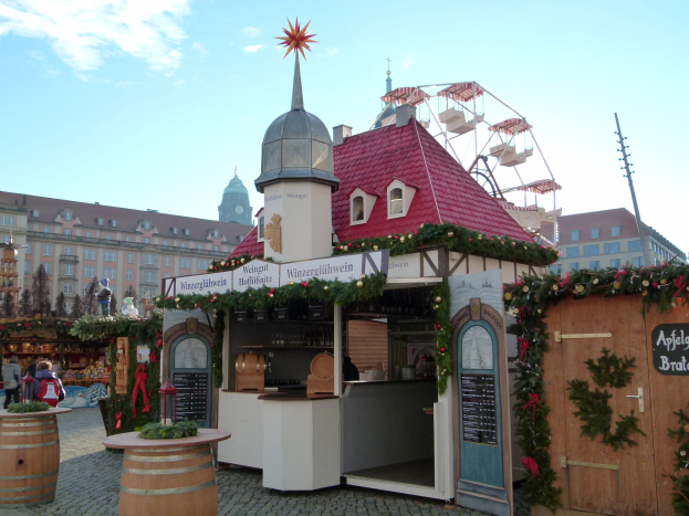 Kleines Gebäude mit rotem Dach und Riesenrad davor, umgeben von Festdekorationen und Menschen, mit Gebäuden, Bäumen und wolkenverhangenem Himmel im Hintergrund.