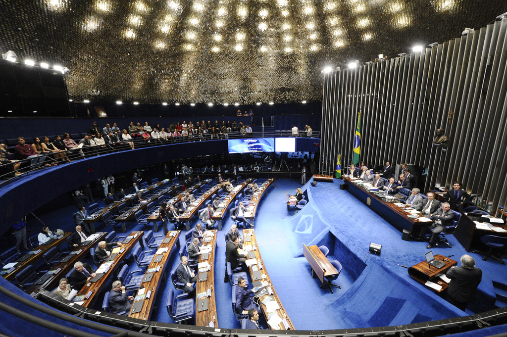 Eine Konferenzbühne mit sitzenden Personen an einem Tisch mit Papieren und Gegenständen, flankiert von Fahnen und einer Skulptur im Hintergrund, mit Sitzplätzen im Vordergrund und stehenden Personen hinter einem Geländer mit Bildschirmen und Deckenbeleuchtung.