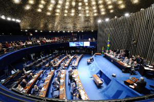 Eine Konferenzbühne mit sitzenden Personen an einem Tisch mit Papieren und Gegenständen, flankiert von Fahnen und einer Skulptur im Hintergrund, mit Sitzplätzen im Vordergrund und stehenden Personen hinter einem Geländer mit Bildschirmen und Deckenbeleuchtung.