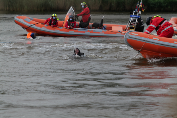 Eine Gruppe von Menschen in einem aufblasbaren Boot auf einem Fluss, mit zwei Personen im Wasser im Vordergrund und Pflanzen im Hintergrund, alle mit Schwimmwesten und Helmen ausgestattet, was auf einen Rettungseinsatz hinweist.