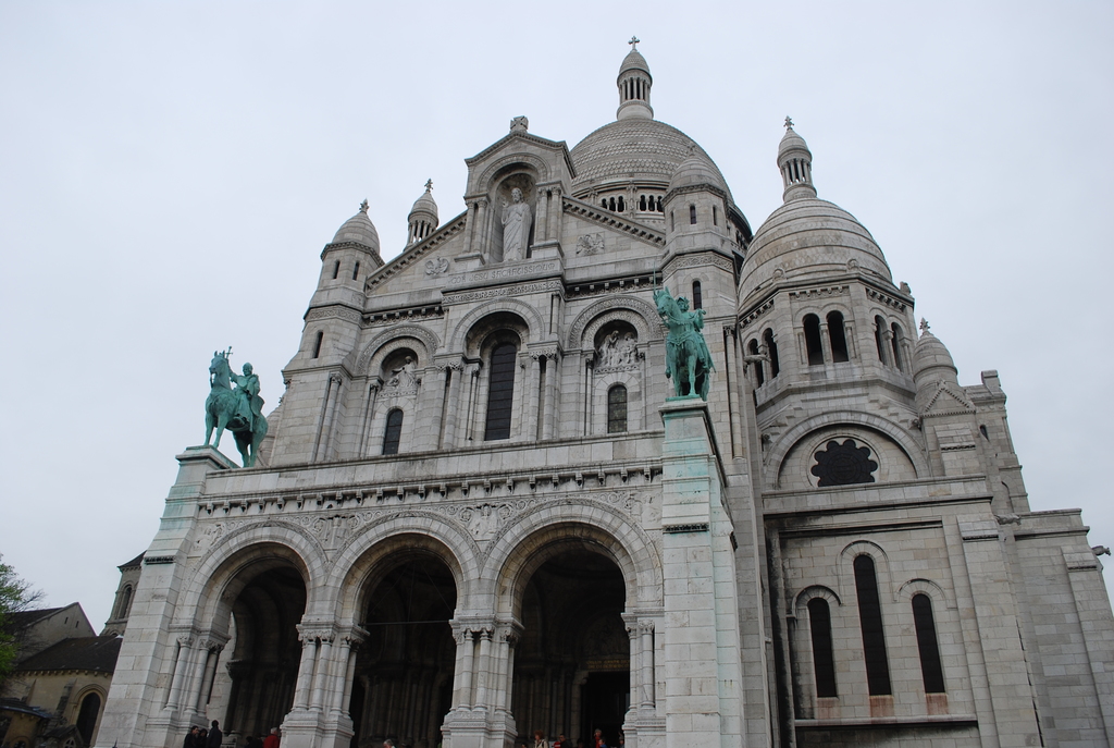 Ein Denkmal steht in der Mitte des Bildes, mit Häusern und einem Baum in der unteren linken Ecke.
