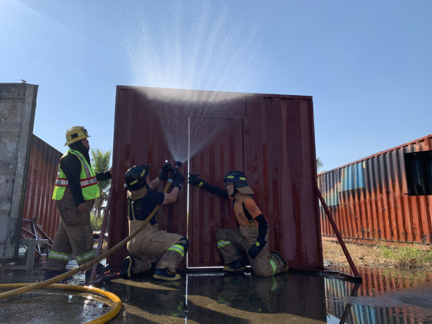 Feuerwehrleute in Helmen und Handschuhen sprühen Wasser aus einem Schlauch auf einen Container, mit Bäumen und einem klaren blauen Himmel im Hintergrund, einem Rohr links und einem Metallcontainer rechts.