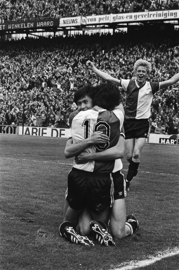 Drei Fussballspieler umarmen sich auf dem Boden nach einem Tor, mit jubelnder Menge und schwenkenden Fahnen in einem schwarz-weiss Stadion-Setting.
