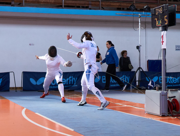 Zwei Frauen in weißen Fechtanzügen und Helmen kämpfen in einem Turnsaal gegeneinander, während eine Zuschauermenge, eine Kamera auf einem Stativ, eine digitale Anzeigetafel und Banner mit Text im Hintergrund zu sehen sind.