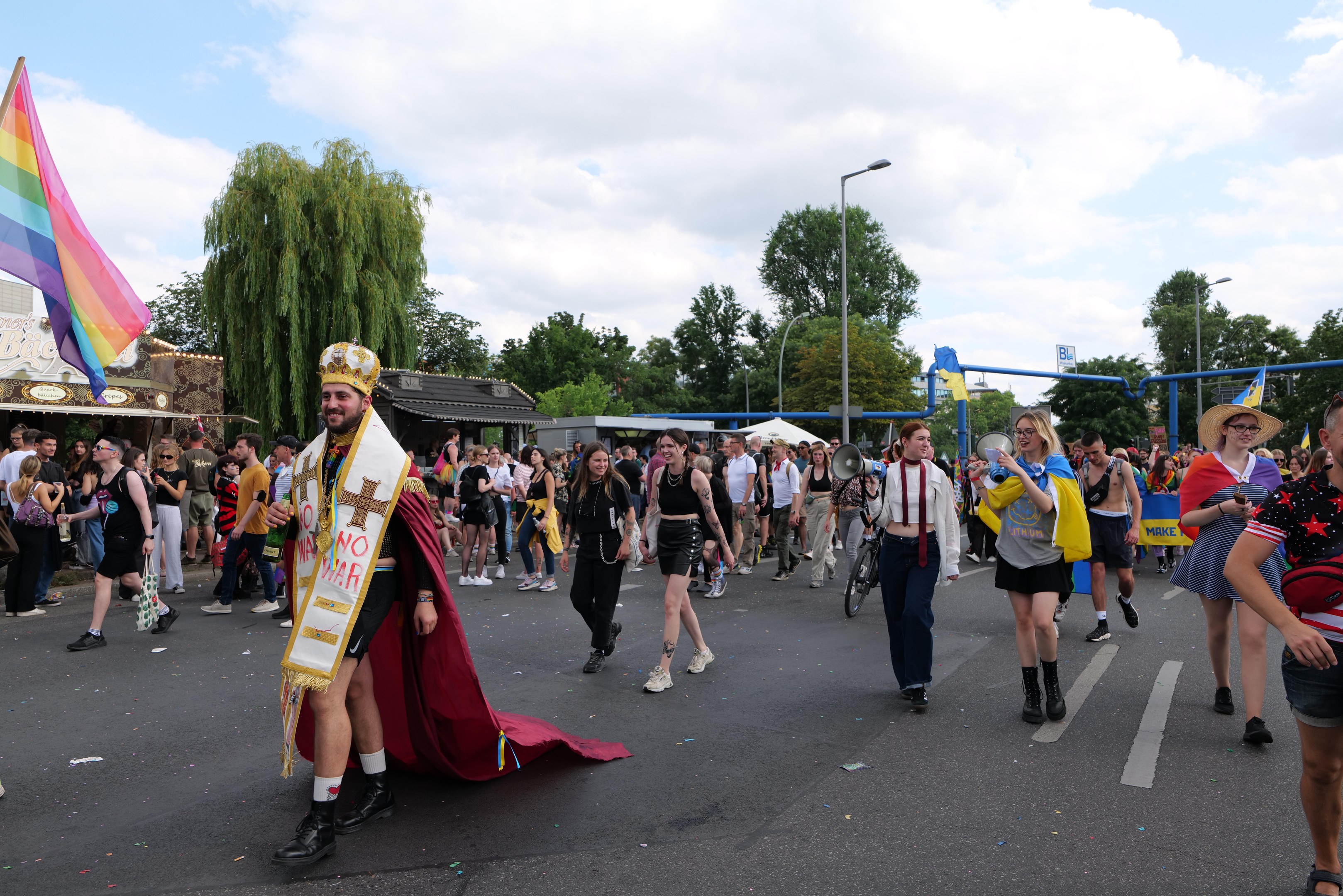 Eine Gruppe von Menschen bei der 2018er Gay Pride Parade mit einer Regenbogenflagge und Musikinstrumenten, einige tragen Mützen, vor dem Hintergrund von Laternenmasten, Bäumen, Schuppen und einem bewölkten Himmel.