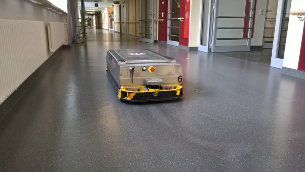 Ein Roboter, der zum Reinigen von Böden entworfen wurde, steht auf dem Boden eines Gebäudes, umgeben von Wänden, Türen, Lampen und anderen Gegenständen.