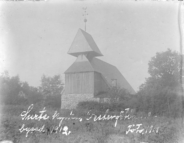 Schwarzes und weißes Foto der Kirche des Heiligen Grabes, das einen Kirchturm, Bäume, Pflanzen und einen bewölkten Himmel zeigt.