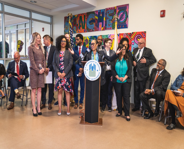 Gruppe von Menschen vor einem Rednerpult mit Mikrofon und einer Tafel mit der Aufschrift "Sancho-Stadtrat tritt dem Stadtrat bei", mit sitzenden und stehenden Teilnehmern, Flaggen, einem Feuerlöscher und Deckenleuchten im Hintergrund.
