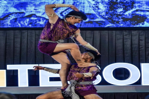 Zwei Akrobaten treten auf der Bühne auf, der Mann hebt die Frau in lila Kleidern hoch, während das Publikum darunter zusieht.