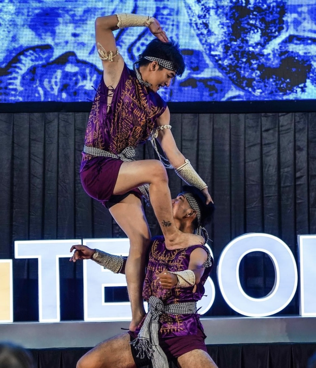 Zwei Akrobaten treten auf der Bühne auf, der Mann hebt die Frau in lila Kleidern hoch, während das Publikum darunter zusieht.