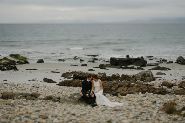Eine Braut und ein Bräutigam sitzen auf einem felsigen Strand, umarmend und küssend mit dem Ozean und dem Himmel im Hintergrund.