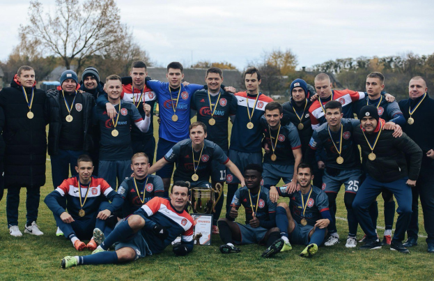 Eine Gruppe von Männern in Medaillen steht auf einem Fußballfeld und hält einen Pokal, umgeben von Bäumen unter einem klaren blauen Himmel.