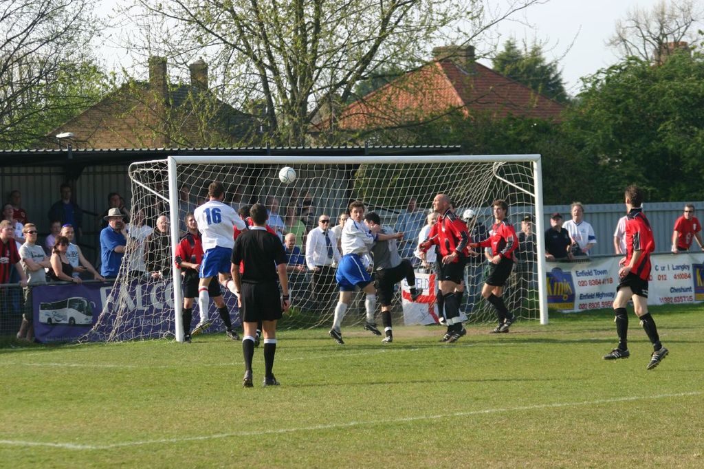 Spieler sind in einem Fußballspiel auf einem Feld mit einem Tornetz beteiligt, während Zuschauer dahinter stehen, mit Bäumen und Häusern im Hintergrund.