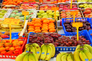 Ein Marktstand mit einer Auswahl an Obst und Gemüse, darunter Bananen, Äpfel, Orangen und Trauben, mit Preisschildern an jedem Artikel.