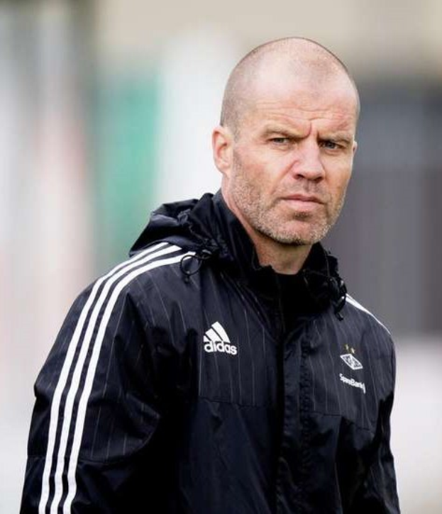 Ein kahlköpfiger Mann in einer schwarzen Jacke steht auf einem Fußballfeld, mit einer marineblauen Jacke mit weißen Streifen und einem weißen Kragen, die Hände verschränkt und einen ernsten Gesichtsausdruck.