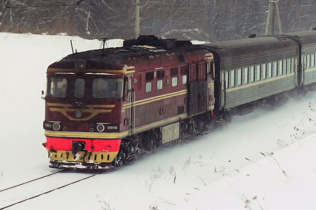 Ein Zug fährt entlang von schneebedeckten Schienen mit Strommasten und Bäumen im Hintergrund, umgeben von einer Schneedecke.