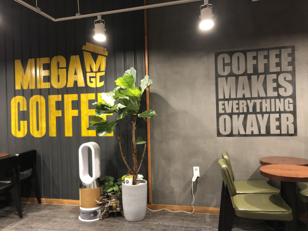 Ein Kaffeehaus-Interieur mit Tischen, Stühlen, einem "Mega Coffee"-Schild an der Wand, einer Bodenstehmaschine, Deckenleuchten und einem Topfpflanze.