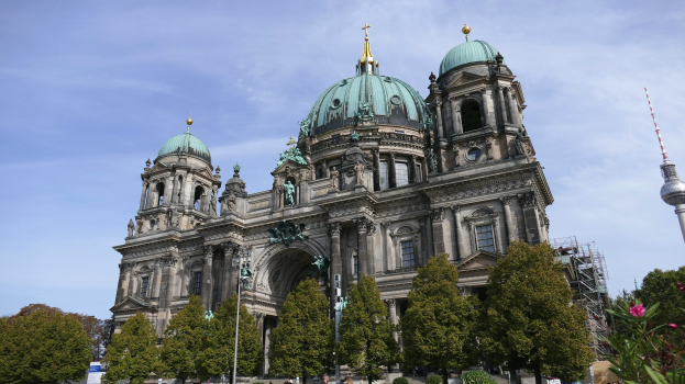 Großes, prunkvolles Berliner Dom-Gebäude in Berlin, Deutschland, mit Statuen, Säulen und gebogenen Fenstern, vor einem bewölkten Himmel, mit Passanten, Bäumen und einem Turm im Vordergrund.