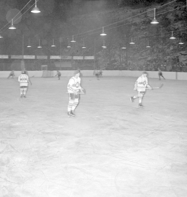 Schwarzes und weißes Foto einer Gruppe von Menschen, die Hockey auf einem Eisstadion unter Deckenlampen spielen, umgeben von einer Wand.