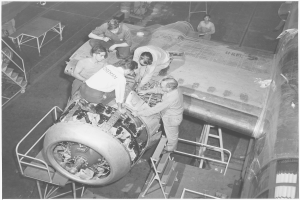 Eine Gruppe von Männern, die an einem Strahltriebwerk in einer schwarzen und weißen Fabrikumgebung arbeiten, mit Leitern, Tischen und anderen Gegenständen im Hintergrund.