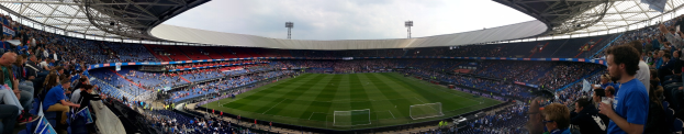 Ein großes Stadion voller Zuschauer, die ein Füballerpiel beobachten, einige halten Kameras, mit einem Netz über das Feld und Laternenmästen gegen einen klaren blauen Himmel.