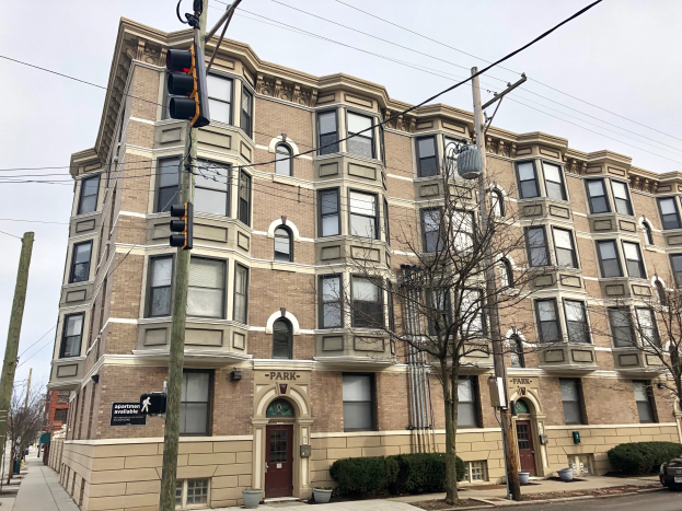 Großes Wohnhaus an einer Straßenecke mit der Aufschrift "Park Apartments for Rent in Chicago, IL", Bäumen, Topfpflanzen, Strommasten, einer Ampel, einem Auto und einem bewölkten Himmel.