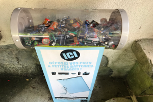 Ein Mülltonnen voller Batterien steht auf einem Bürgersteig, mit einem Plakat mit Text an der Wand dahinter und Treppen auf der rechten Seite.