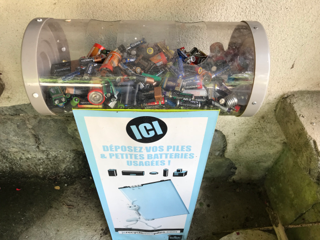 Ein Mülltonnen voller Batterien steht auf einem Bürgersteig, mit einem Plakat mit Text an der Wand dahinter und Treppen auf der rechten Seite.