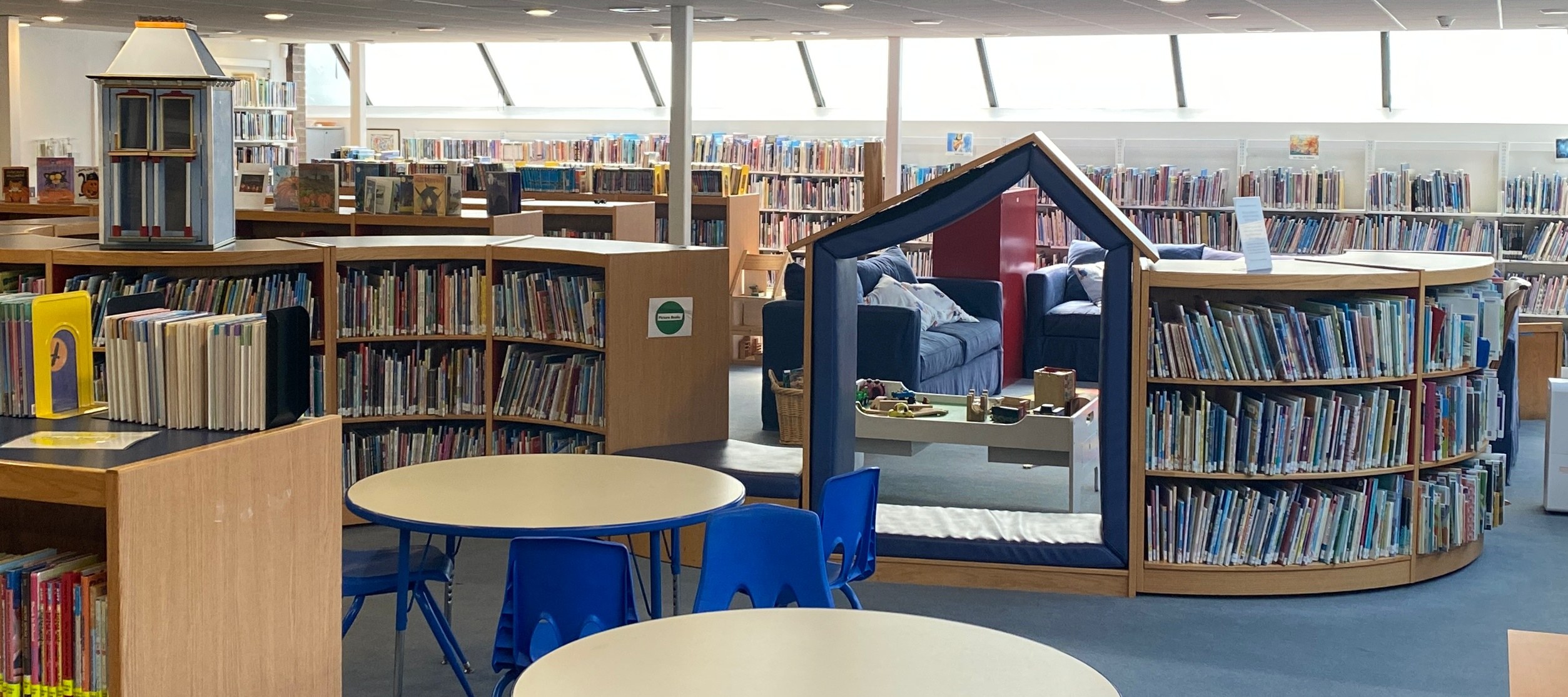 Eine Bibliothek mit Bücherregalen, Tischen, Stühlen, Sofas mit Kissen, Deckenleuchten und einem Pfosten.
