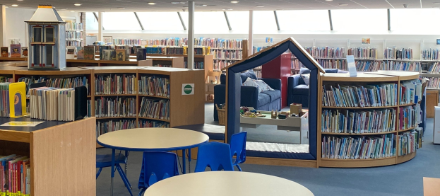 Eine Bibliothek mit Bücherregalen, Tischen, Stühlen, Sofas mit Kissen, Deckenleuchten und einem Pfosten.