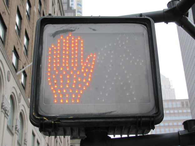Eine Ampel mit roter Hand in der Mitte einer Stadt, umgeben von Gebäuden auf beiden Seiten und einem klaren Himmel darüber.