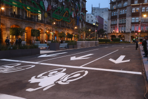 Eine Stadtstraße mit einem Fahrradweg, Gebäuden, Straßenlaternen, Verkehrsschildern, Menschen auf dem Gehweg, Bänken, Pflanzen, Bäumen, dekorativen Gegenständen und einem klaren blauen Himmel.