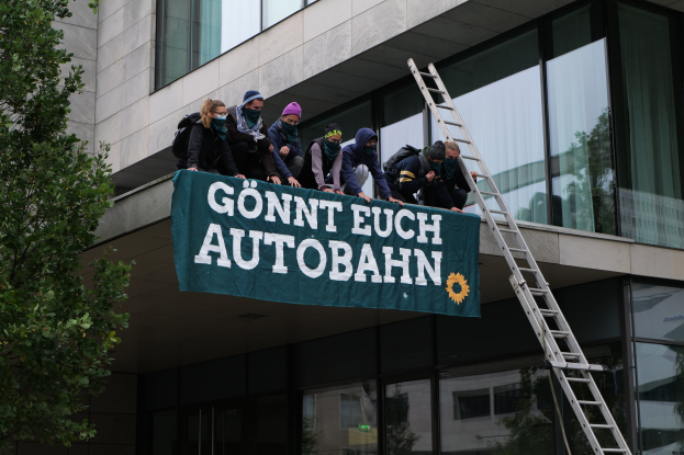 Eine Gruppe von Menschen auf dem Dach eines Gebäudes stehend, eine Fahne haltend, mit einem Baum links und Glasfenstern im Hintergrund, wahrscheinlich protestierend im Support der deutschen Regierung.