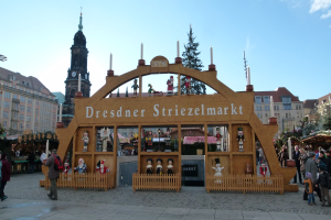 Ein lebendiger Weihnachtsmarkt in Dresden, Deutschland, mit Menschen, die herumlaufen, einige mit Taschen, und einem Bogen mit Text darauf, vor einer Kulisse aus Bäumen, Gebäuden mit Fenstern und einem bewölkten Himmel.