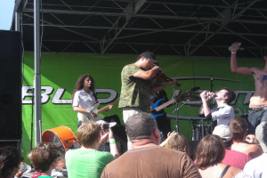 Eine Gruppe von Musikern performs auf der Buhne mit verschiedenen Instrumenten, waehrend das Publikum vor einem Podium steht und die Musik genießt. Ein grosser schwarzer Lautsprecher ist links zu sehen, und ein grunes Banner ist im Hintergrund sichtbar.