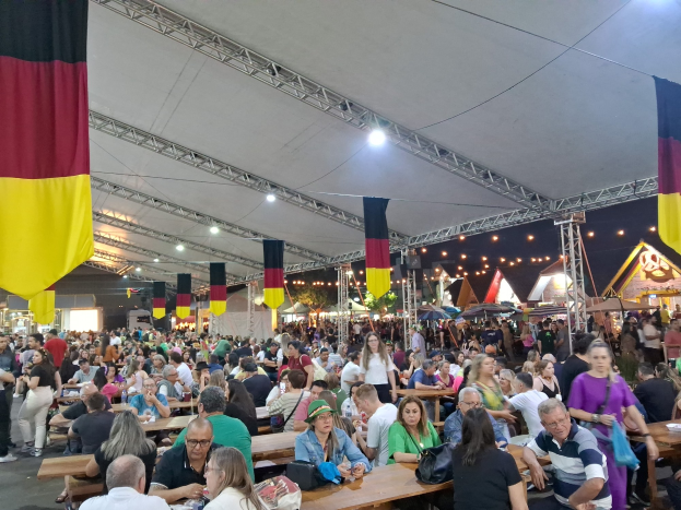 Eine große Gruppe von Menschen sitzt an Tischen unter einem Zelt auf dem Oktoberfest in München, Deutschland, einige stehen und Taschen liegen auf den Tischen, umgeben von festlichen Zelten, Lichtern und Fahnen.