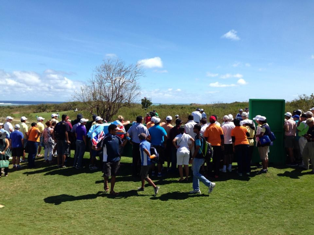 Gruppe von Menschen auf einem grünen Feld bei einem Golfturnier, mit Bäumen, Pflanzen und einem Gewässer im Hintergrund.
