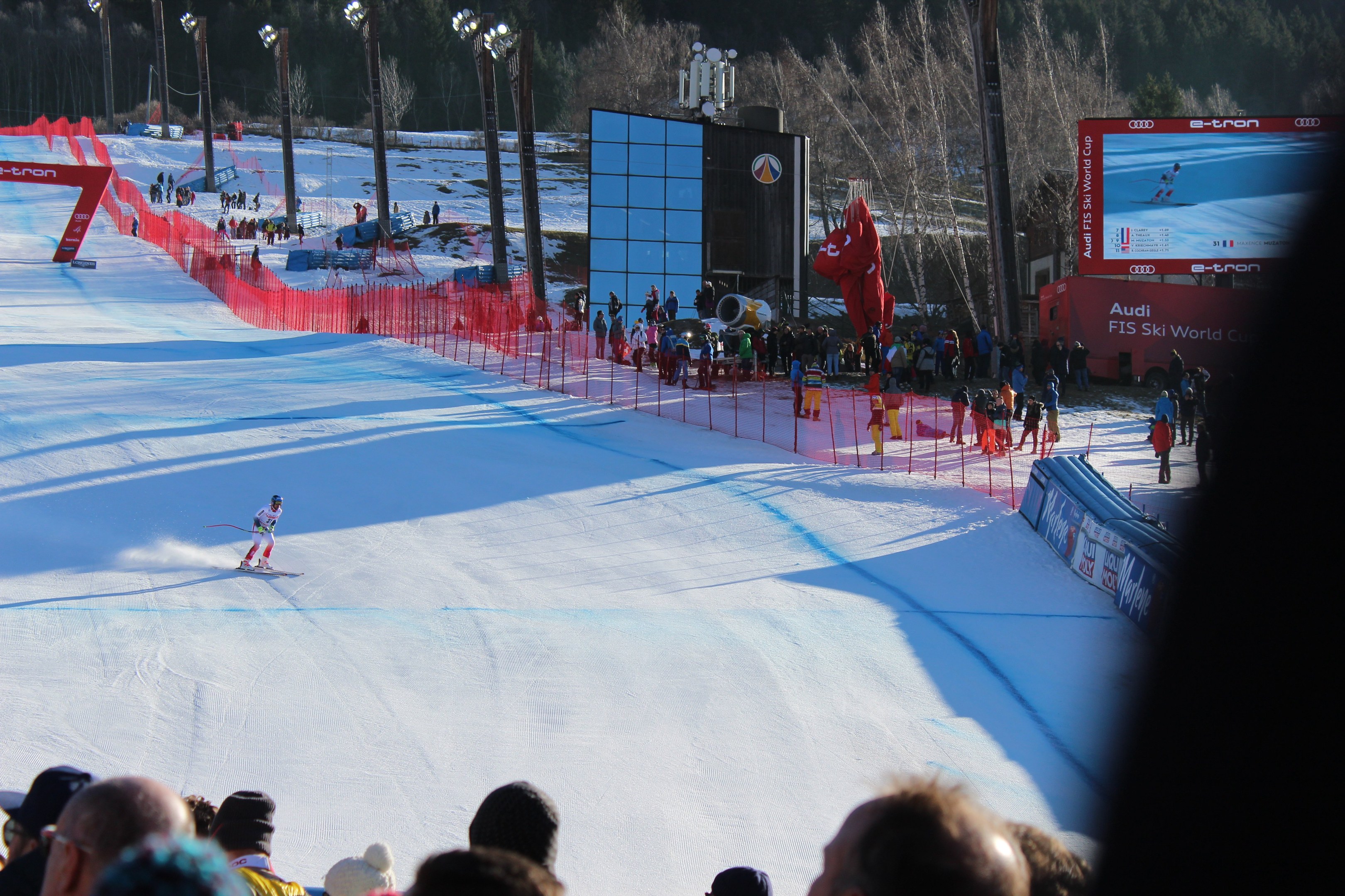 Eine Person fährt Ski auf einer schneebedeckten Piste vor einer Zuschauermenge, mit einem Zaun, Bäumen, Laternen und einem Bildschirm mit Text auf der rechten Seite.