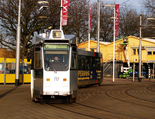 Weißer und blauer Tram fährt auf einer Stadtstraße mit einem Passagier im Inneren, umgeben von städtischer Infrastruktur und Grünfläche unter einem klaren Himmel.