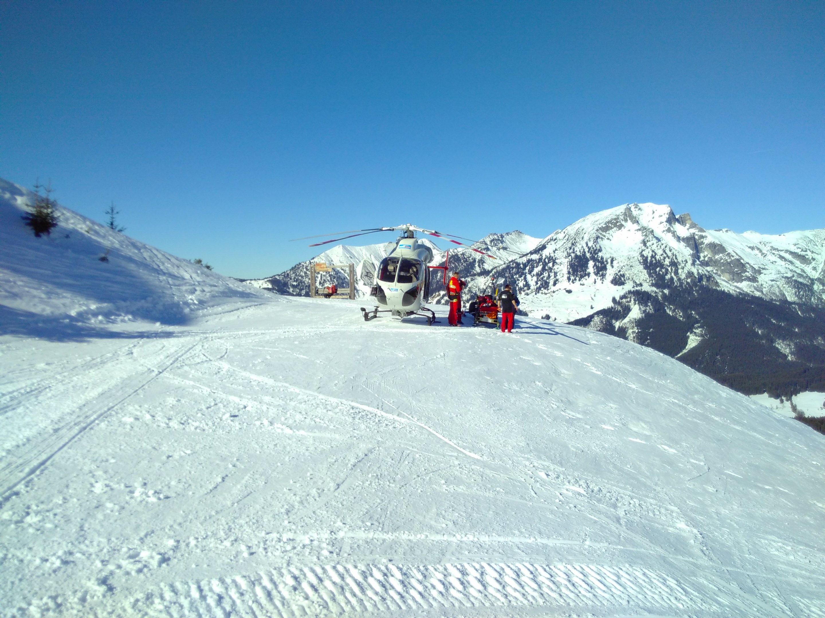 Hubschrauber auf schneebedecktem Berg mit wenigen Menschen in der N├Ąhe, schneebedeckte Berge im Hintergrund bei klarem Himmel.