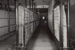 Ein Schwarz-Weiß-Foto eines langen Gangs in einem Rechenzentrum, das Metallobjekte, Rohre und Deckenleuchten zeigt, mit zahlreichen Servern und Regalen voller Daten.