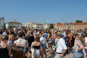 Eine große Menschenmenge steht vor einem Gebäude mit Fenstern, Laternenmasten, Flaggen und Bäumen, einige halten Kameras, was auf eine Protest- oder Demonstrationsveranstaltung unter einem bewölkten Himmel hinweist.