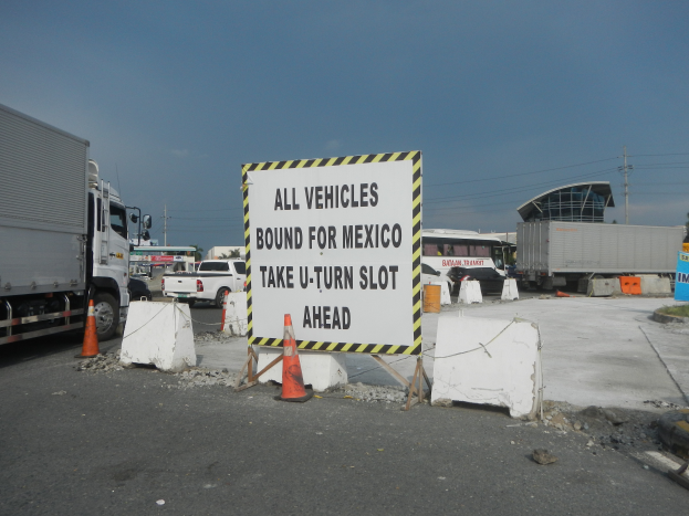 Ein Lastwagen auf einem Parkplatz neben einem Schild mit der Aufschrift "Alle Fahrzeuge nach Mexiko", mit verstreuten Gegenständen, Gebäuden, Bäumen, Polen und einem klaren blauen Himmel im Hintergrund.