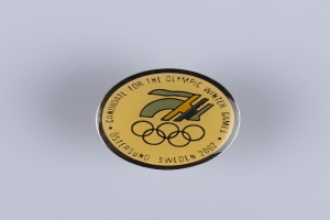 Ein rundes Abzeichen mit schwarzem Hintergrund und goldfarbenem Olympische Winterspiele-Logo in der Mitte, das die Schrift "Olympische Winterspiele" in fetter weißer Schrift auf einer weißen Fläche zeigt.