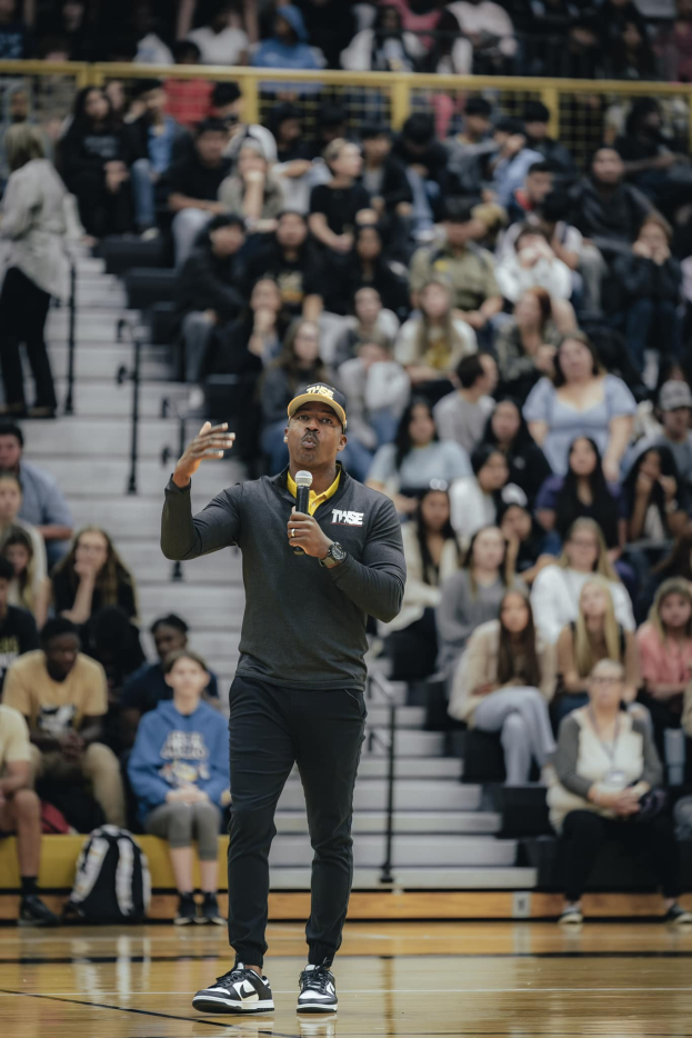 Ein Mann in einer Mütze und mit Mikrofon steht auf einem Basketballfeld, während Zuschauer auf der Treppe hinter ihm sitzen und Taschen auf dem Boden liegen.