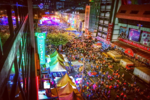 Eine volle KarnevalsSzene mit Zelten, Geschäften, Gebäuden und Bannern, beleuchtet von Lichtern im Hintergrund.