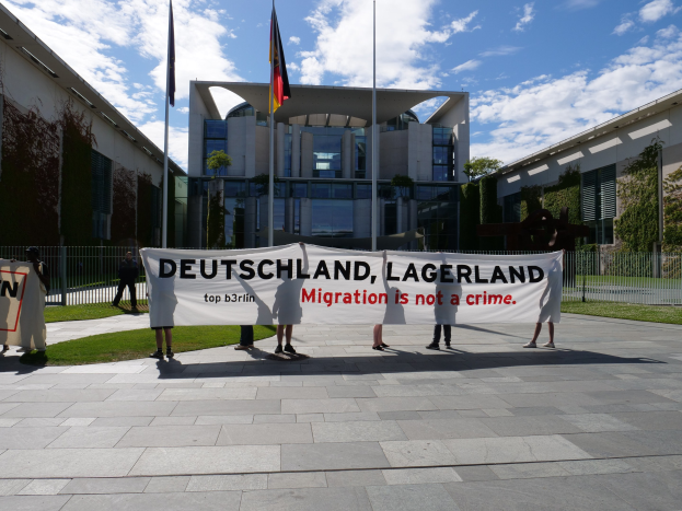 Gruppe von Menschen mit einem Banner mit der Aufschrift "Deutschland, Lagerland Migration ist kein Verbrechen" vor einem Gebäude mit Fenstern, umgeben von Gras, einem Metallzaun, Pflanzen, Bäumen und Fahnen mit Stangen unter einem bewölkten Himmel.