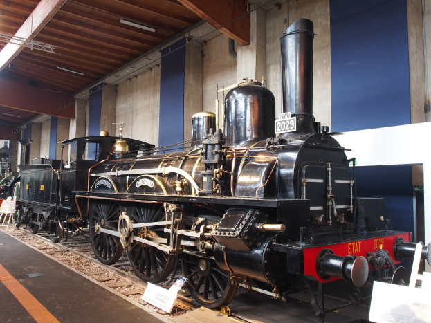 Eine große, alte Dampflokomotive in einem Museums-Setting umgeben von Eisenbahnschienen, Informations-Tafeln und anderen Ausstellungsstücken, mit einer Wand und beleuchteter Decke im Hintergrund.