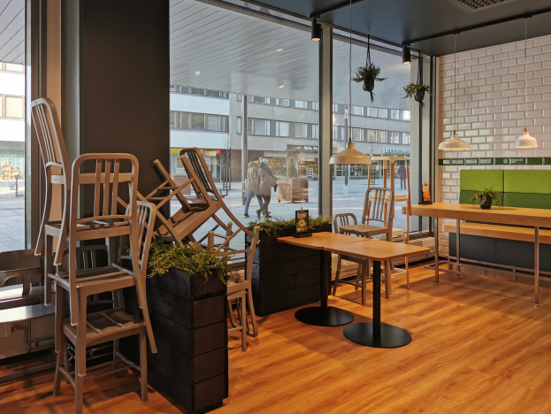 Ein Restaurantinnenraum mit hölzernen Tischen und Stühlen in der Nähe eines großen Fensters, das auf eine Berliner Straße mit Passanten, Gebäuden und urbanen Elementen hinausgeht.