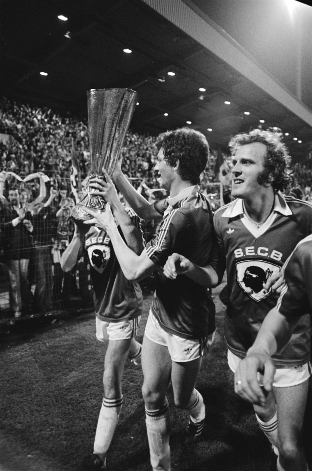 Schwarzes und weißes Bild von Fußballspielern, die einen Pokal auf einem Feld halten und ihren FA-Cup-Finalsieg feiern, mit Zuschauern hinter einem Zaun unter Stadionbeleuchtung.